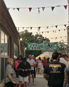 Festival-goers walk beneath a banner reading “Welcome to the Feast of the Assumption,” with tents and lights lining the street in Little Italy.