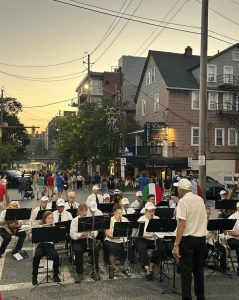 A community band performs outdoors at sunset under the direction of a conductor, with festival crowds walking the Little Italy streets behind them.