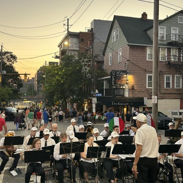 A community band performs outdoors at sunset under the direction of a conductor, with festival crowds walking the Little Italy streets behind them.