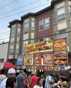 Crowds line up at a food stand decorated with bright signs advertising Italian sausage sandwiches, meatball subs, and other festival favorites.