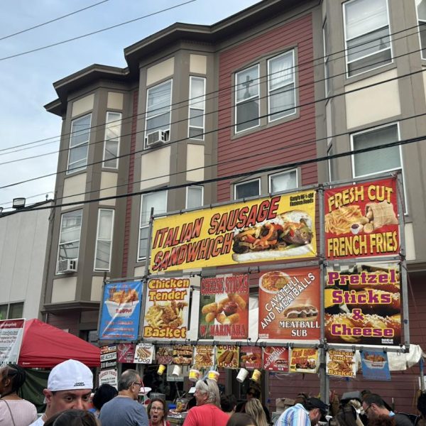 Crowds line up at a food stand decorated with bright signs advertising Italian sausage sandwiches, meatball subs, and other festival favorites.