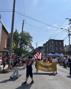 Parade participants march down Mayfield Road carrying Italian and American flags alongside a gold ICCA banner, with spectators filling the sidewalks.