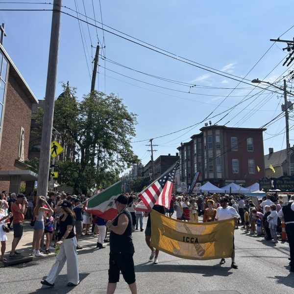 Parade participants march down Mayfield Road carrying Italian and American flags alongside a gold ICCA banner, with spectators filling the sidewalks.