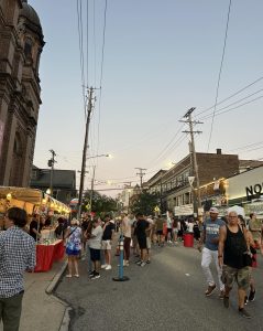 Festival-goers stroll past food stands and vendor tents along Mayfield Road as the evening sky sets over Little Italy.