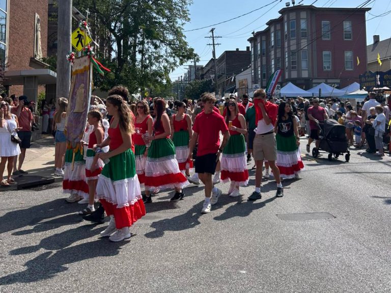 Parade participants dressed in red, white, and green skirts and shirts march down Mayfield Road as crowds look on during the Feast of the Assumption.