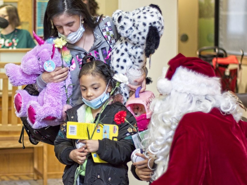 A young girl holding art supplies and a red carnation, accompanied by a woman carrying stuffed animals, meets Santa Claus during a holiday gift event.