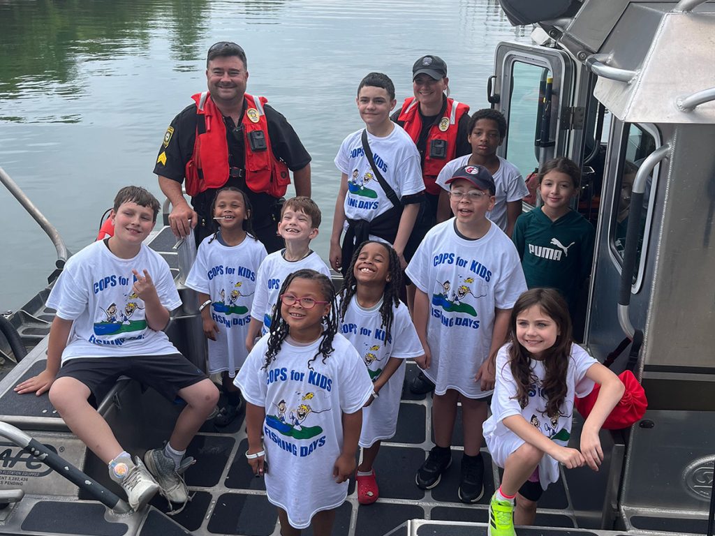 Officers and children in matching “Cops for Kids Fishing Days” shirts gather on a charter fishing boat during a community outing.