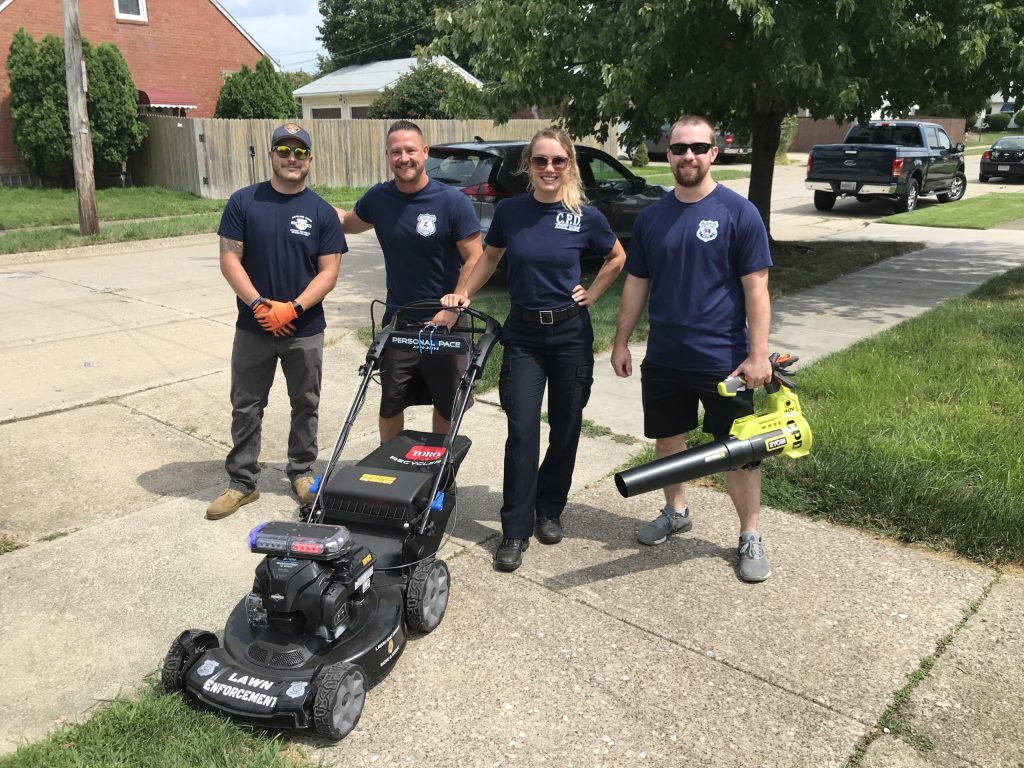Officers stand together outdoors with a lawnmower and leaf blower during a neighborhood cleanup project.