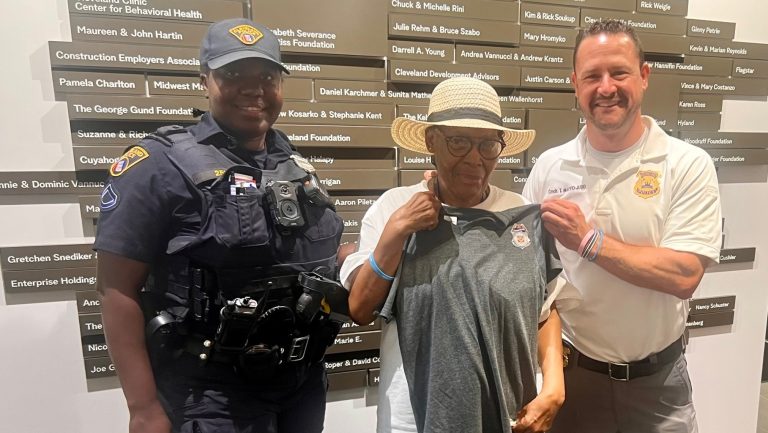 Second District officer and commander present a Cleveland Police T-shirt and badge to a smiling senior resident inside the May Dugan Center.
