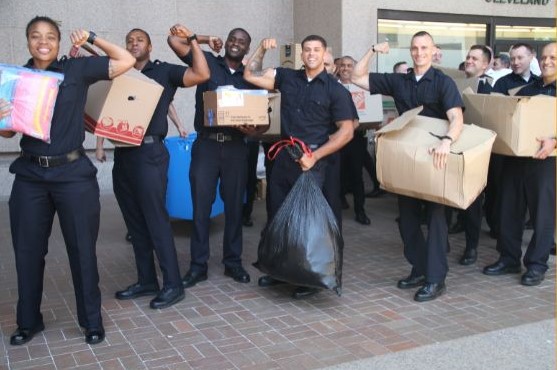 Smiling Cleveland Police Academy recruits pose outside headquarters holding boxes and bags of donations, flexing their arms to celebrate the relief drive for Rainbow Terrace families.