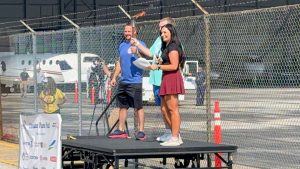 An emcee holds a lit torch on a small stage during the opening ceremony, with aircraft and event staff visible behind a chain-link fence.