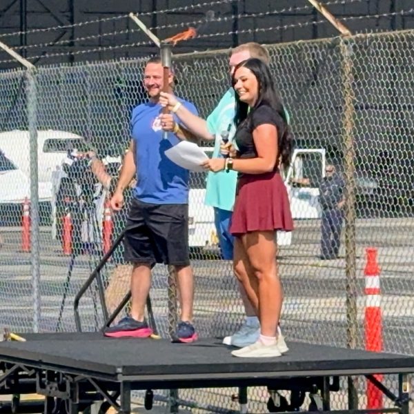 An emcee holds a lit torch on a small stage during the opening ceremony, with aircraft and event staff visible behind a chain-link fence.