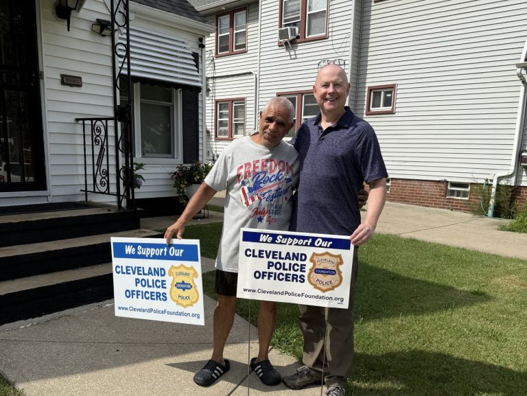 Mr. Pena stands outside his home holding a “Support Our Police Officers” yard sign alongside a supporter, both smiling on the sidewalk.