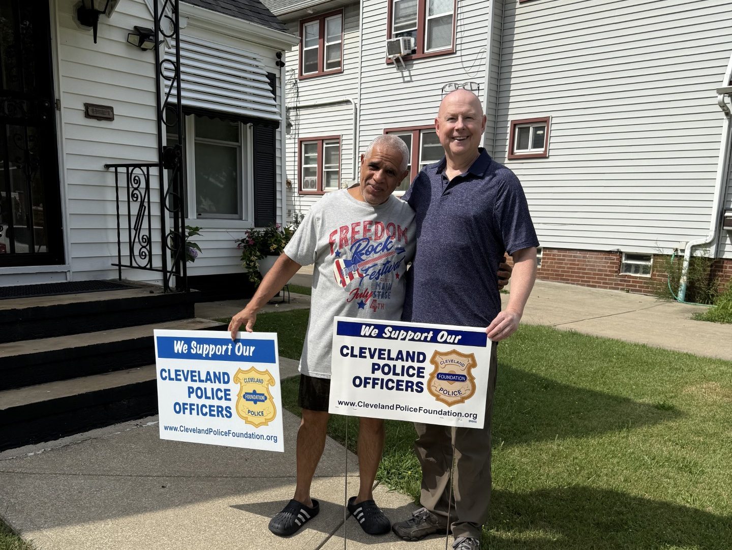 Mr. Pena stands outside his home holding a “Support Our Police Officers” yard sign alongside a supporter, both smiling on the sidewalk.