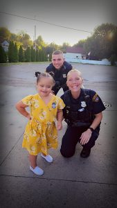 Two Cleveland officers kneel beside a young girl in a yellow dress outdoors, all smiling for the camera.