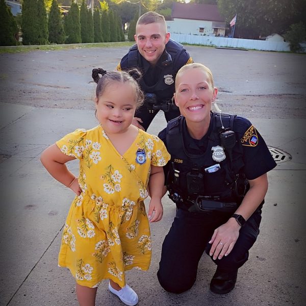 Two Cleveland officers kneel beside a young girl in a yellow dress outdoors, all smiling for the camera.