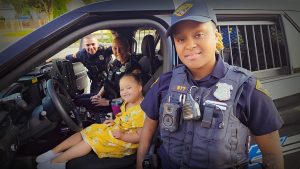 A young girl in a yellow dress sits smiling in the driver’s seat of a Cleveland Police cruiser with two officers inside and another standing by the door.