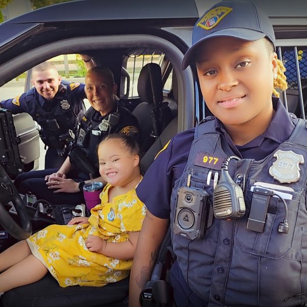 A young girl in a yellow dress sits smiling in the driver’s seat of a Cleveland Police cruiser with two officers inside and another standing by the door.