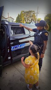 A Cleveland officer and a young girl in a yellow dress point together toward the back of a police cruiser.