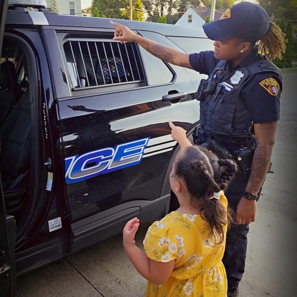 A Cleveland officer and a young girl in a yellow dress point together toward the back of a police cruiser.