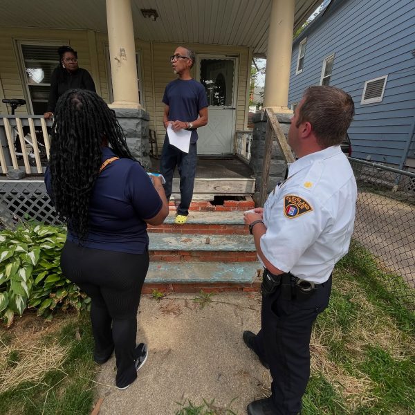 A Cleveland Police officer and a city staff member stand on the sidewalk speaking with a resident holding papers on his front porch, while another woman looks on from the porch railing.