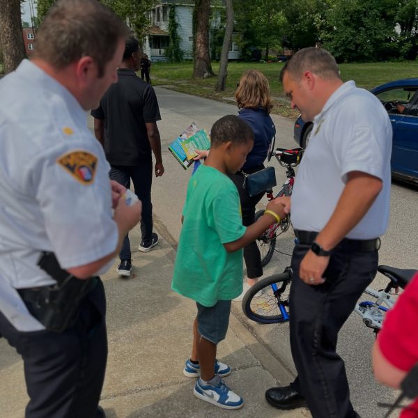 Two Cleveland Police officers interact with a young boy in a green shirt standing beside his bicycle, handing him materials during the neighborhood safety walk.