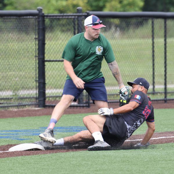 Player in a green shirt attempts to tag out a sliding runner in a black Cleveland Police softball jersey at third base.
