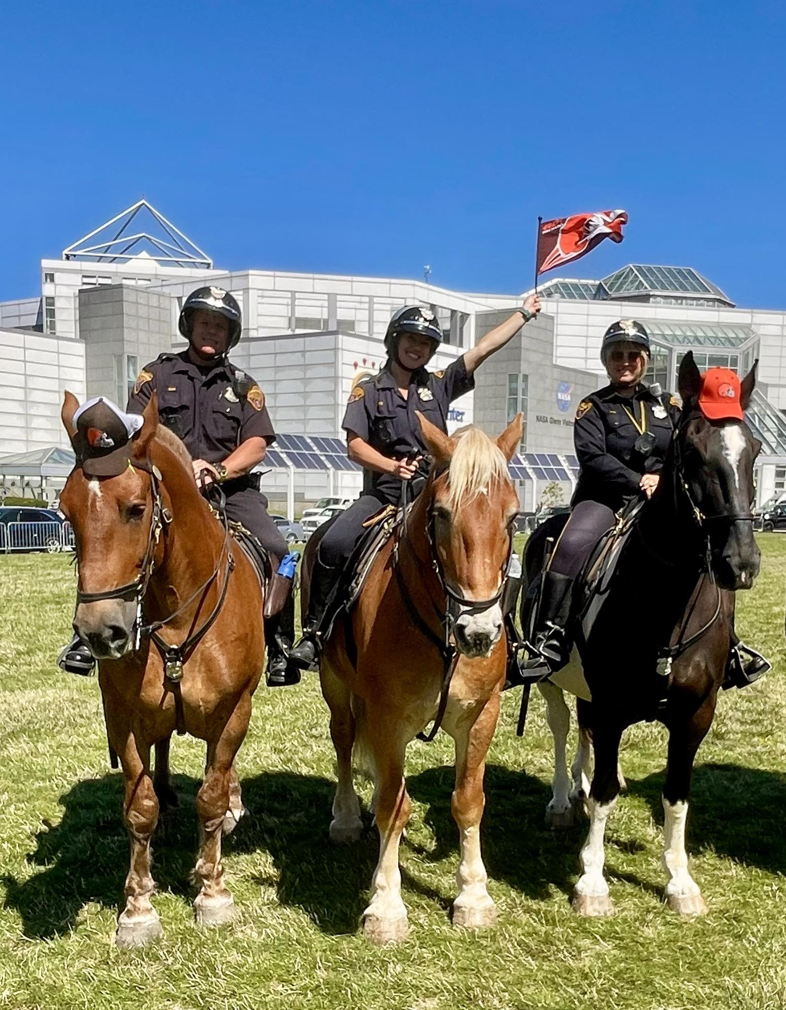 Three Cleveland Police Mounted Unit officers on horseback outside Cleveland Browns Stadium, with one officer holding a Browns flag while the horses stand on the grass.