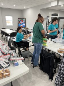 Volunteers in teal shirts prepare donation bags with snacks and supplies inside a community space.