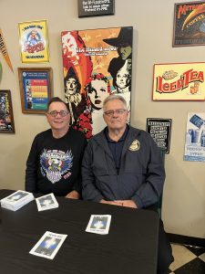 Two men seated at a table with brochures, one wearing a Support Our First Responders shirt and the other a jacket with a police badge patch, with colorful posters behind them.