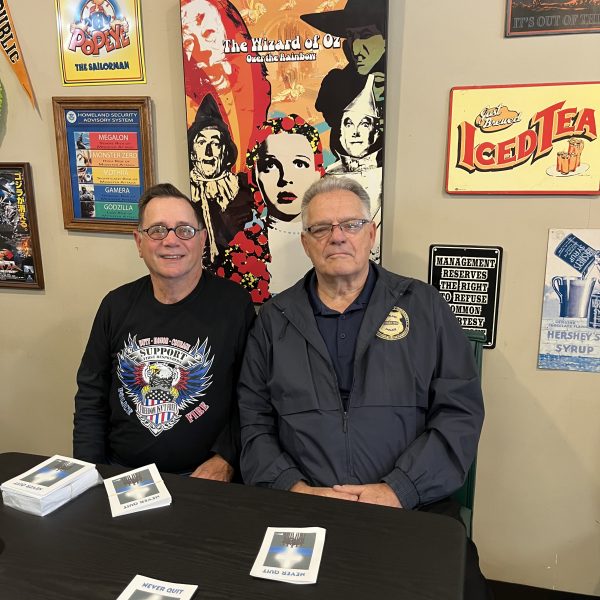 Two men seated at a table with brochures, one wearing a Support Our First Responders shirt and the other a jacket with a police badge patch, with colorful posters behind them.