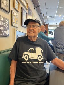 Older man wearing a “Follow me to Savor the Moment” T-shirt and cap, seated inside the cafe.