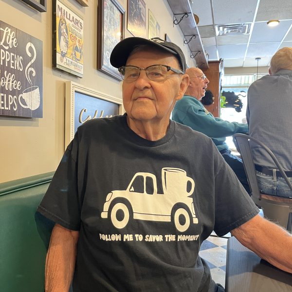 Older man wearing a “Follow me to Savor the Moment” T-shirt and cap, seated inside the cafe.
