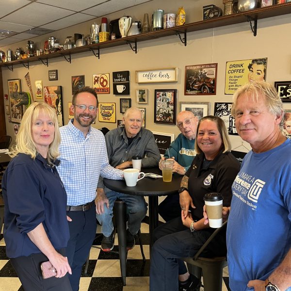 Group of community members and officers standing and seated together with coffee cups inside Savor the Moment cafe.
