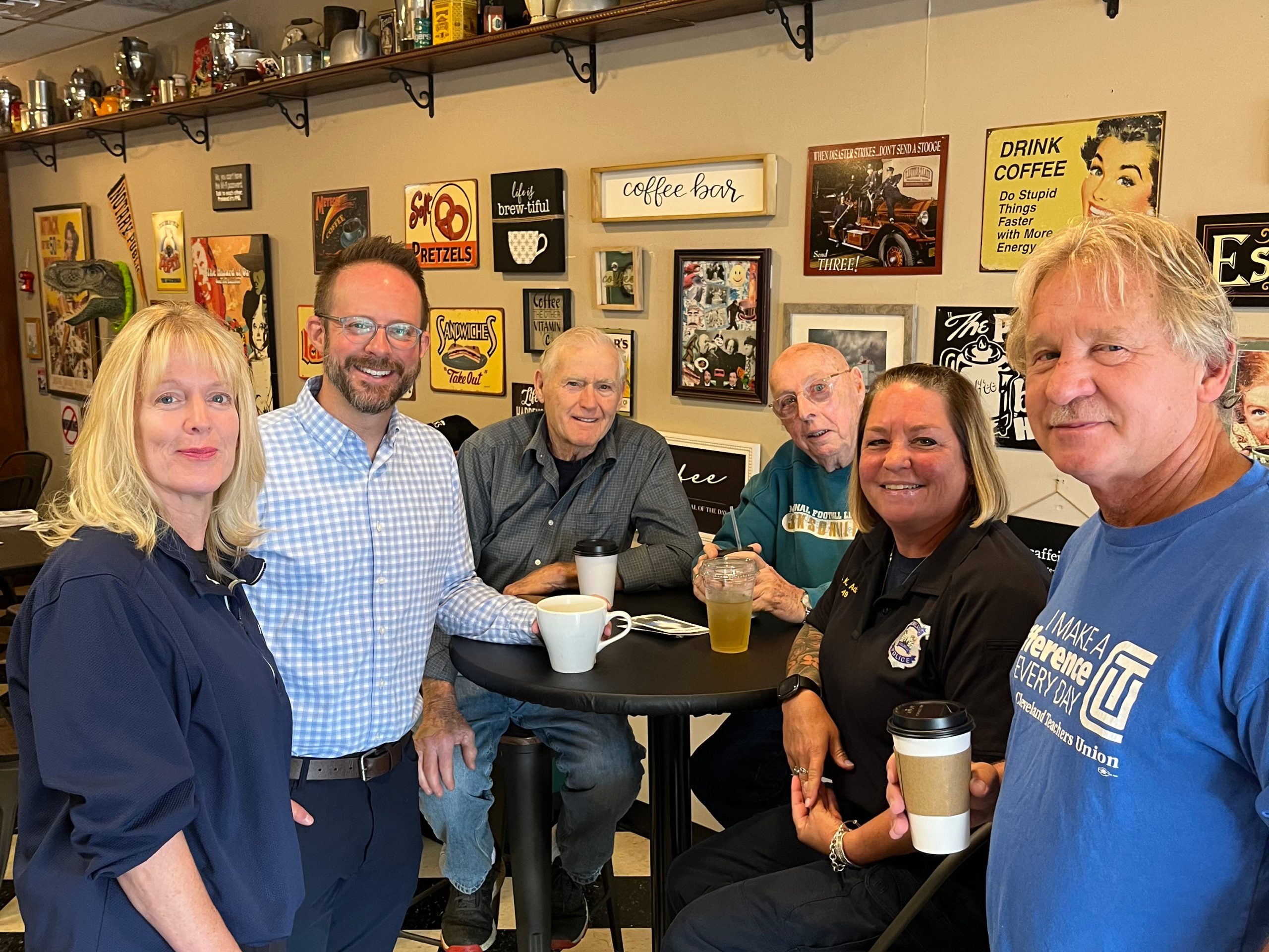 Group of community members and officers standing and seated together with coffee cups inside Savor the Moment cafe.