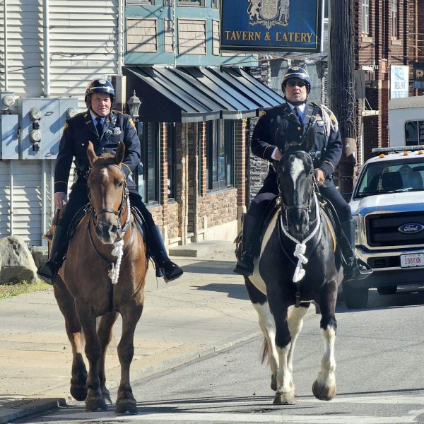 Two mounted officers riding down a city street past storefronts, including a tavern sign.
