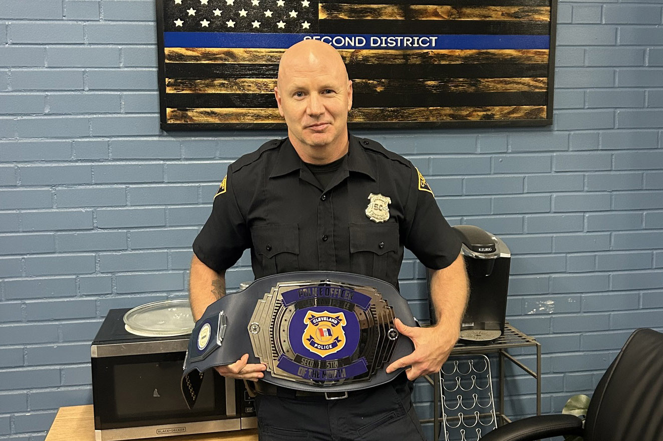 Officer Jason Adkins, Second District Officer of the Month for August 2025, standing in uniform and holding a championship-style belt in front of a thin blue line flag sign.