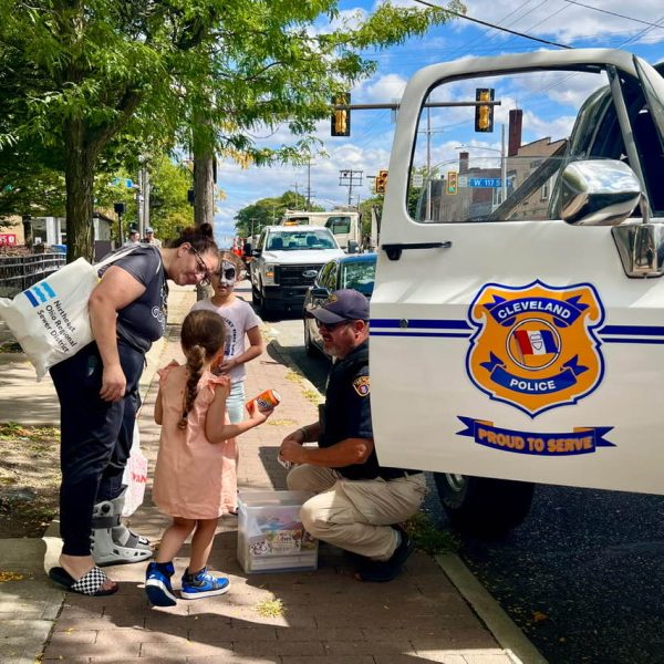 Officer kneeling to hand out treats and coloring books beside the CPD Monster Truck, speaking with children and parents.