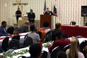 Commander Mark Maguth speaking at the podium, with Harry Boomer and others on stage.