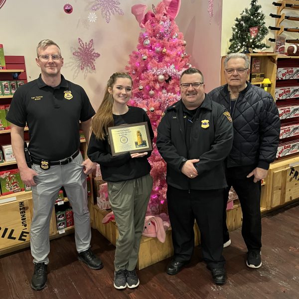 Five adults standing indoors near a pink Christmas tree during an award presentation, with Bob Guttu standing on the far right.