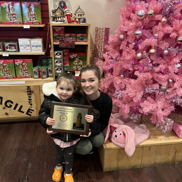 A young child and an adult kneeling together indoors, holding a Community Partner of the Month plaque in front of a pink Christmas tree.