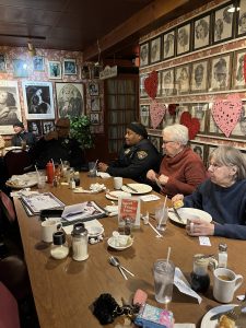 Police officers and community members seated together at a long table inside a restaurant, talking and eating during a Coffee with a Cop event.
