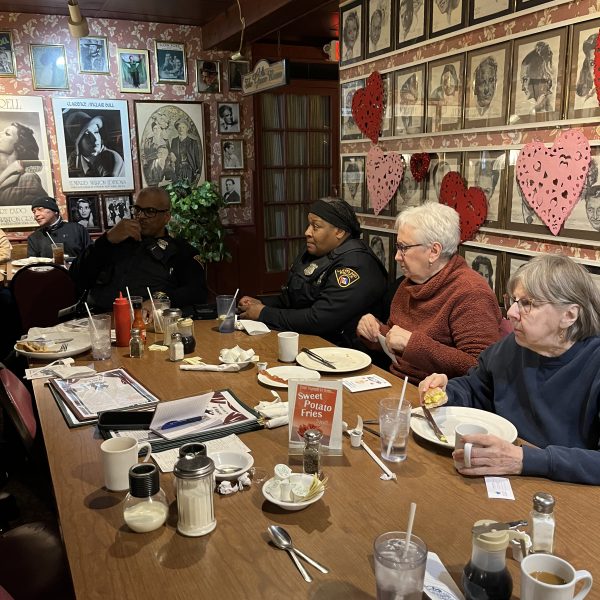 Police officers and community members seated together at a long table inside a restaurant, talking and eating during a Coffee with a Cop event.