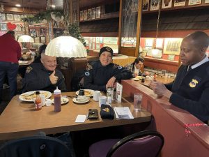 Police officers seated at a restaurant counter and booth, smiling and talking over breakfast during a Coffee with a Cop gathering.