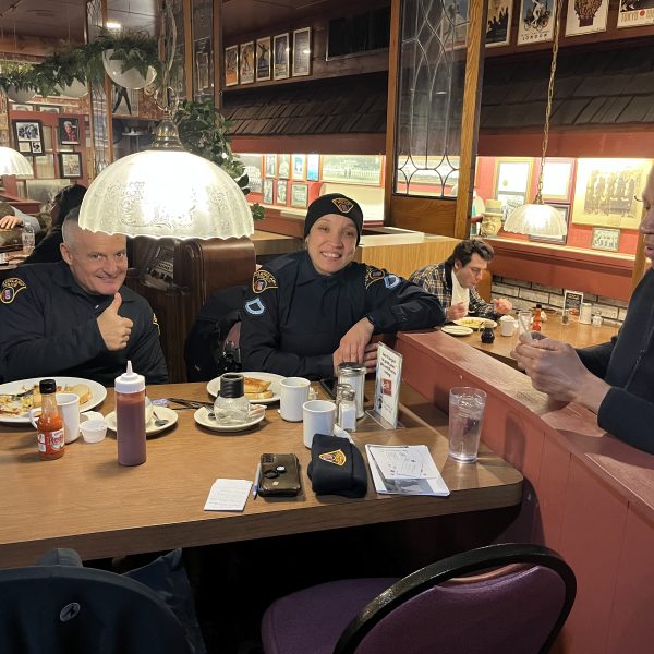 Police officers seated at a restaurant counter and booth, smiling and talking over breakfast during a Coffee with a Cop gathering.
