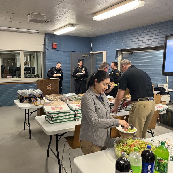 Police officers and staff standing around tables serving themselves food during a group lunch inside a district building.
