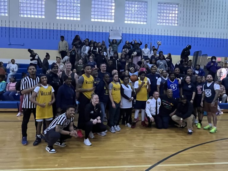 Both teams and officials pose together on the court with a packed crowd of Willson School supporters in the bleachers behind them